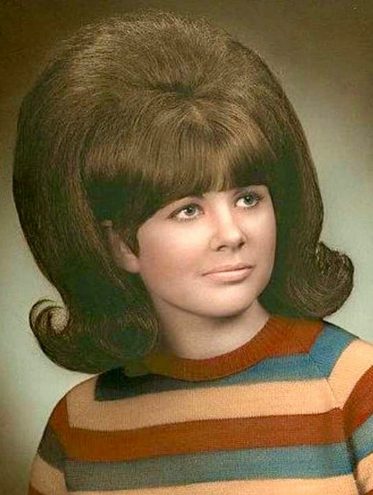 Woman with a voluminous 1960s beehive hairstyle wearing a colorful striped sweater.
