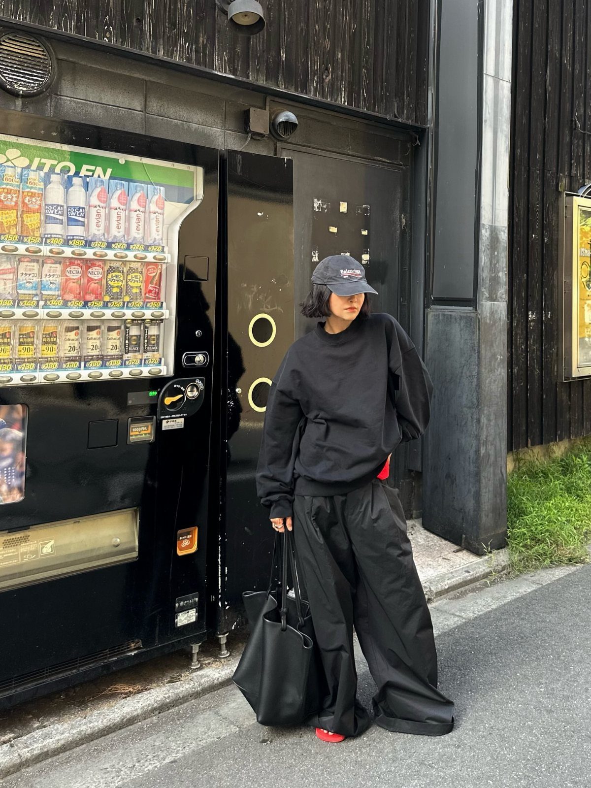 Woman in oversized black sweatshirt and baggy black pants standing by a vending machine.