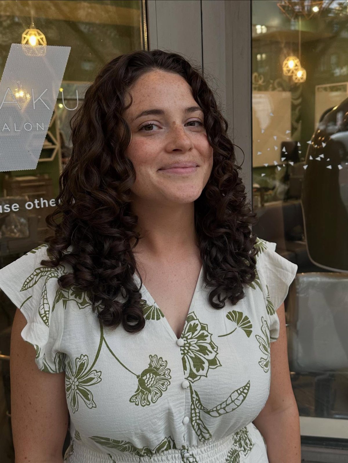 Woman with medium length dark brown curly hair wearing a floral white dress.