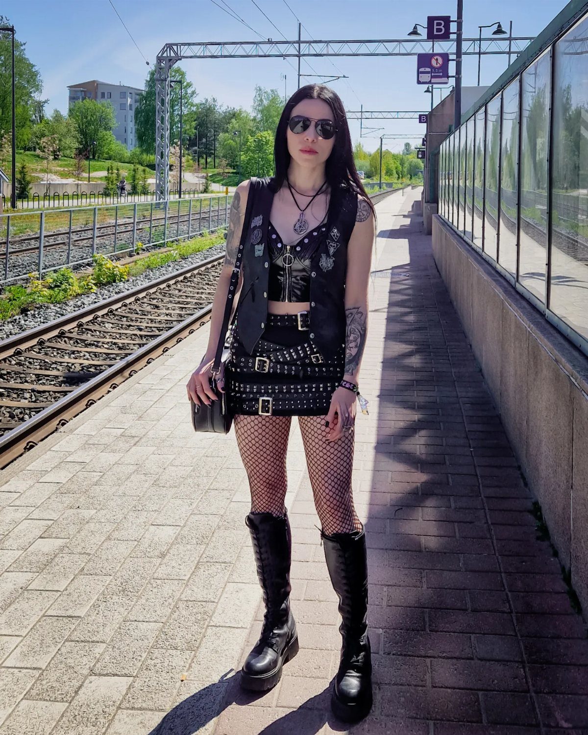 Woman in black leather vest, fishnets, and tall boots posing on a train platform
