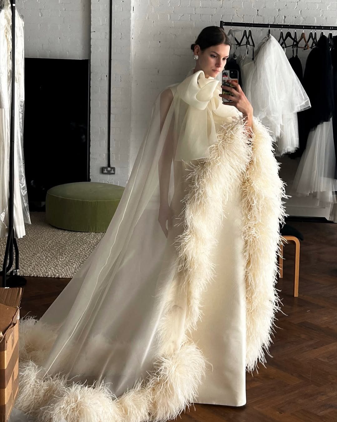 Woman wearing a white bridal gown with a sheer feather-trimmed cape and large bow.