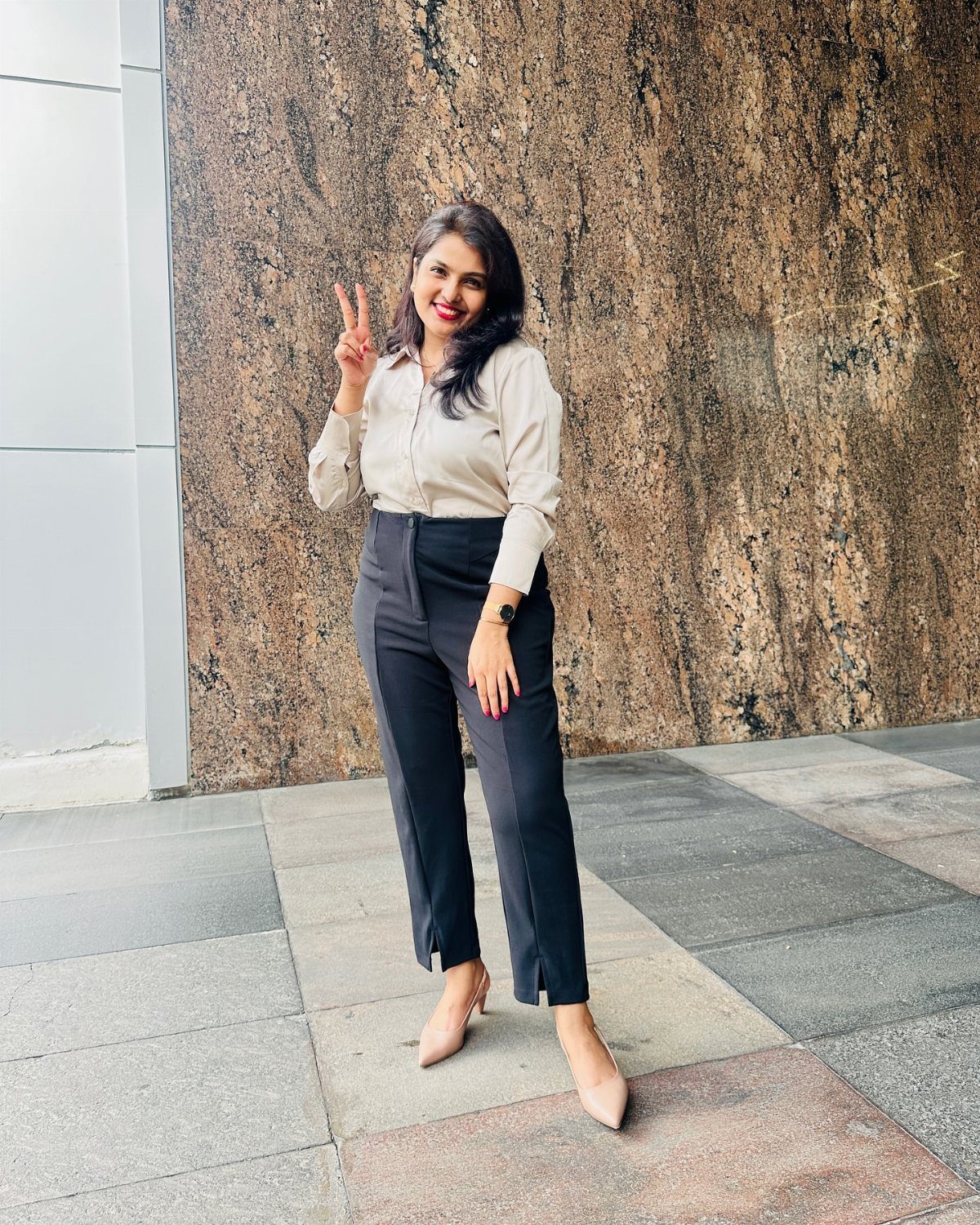 Woman wearing a beige button-down shirt and black trousers standing by a stone wall.